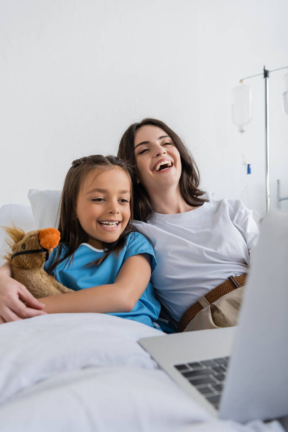 Szczęśliwe dziecko w sukni pacjenta patrząc na laptopa w pobliżu matki i miękkie zabawki na łóżku w szpitalu  - Zdjęcie, obraz
