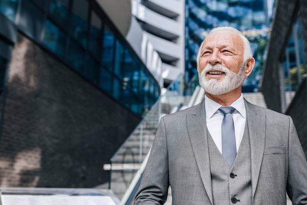 Confident senior businessman with beard looking away. Elderly smiling male entrepreneur is standing against modern office building in the city. He is wearing full suit. - Fotoğraf, Görsel