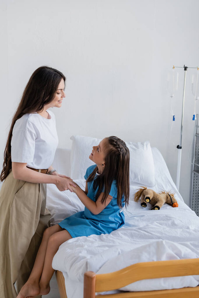 Annenin hasta önlüklü kızının elini tutuşu, klinikteki yatakta.  - Fotoğraf, Görsel