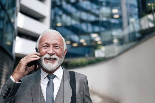 スマートフォンで話し合う先輩ビジネスマンの笑顔のクローズアップ。オフィスビルの反対側に立って袋を持つ高齢男性の専門家。彼は街を旅行中にスーツを着ている. - 写真・画像