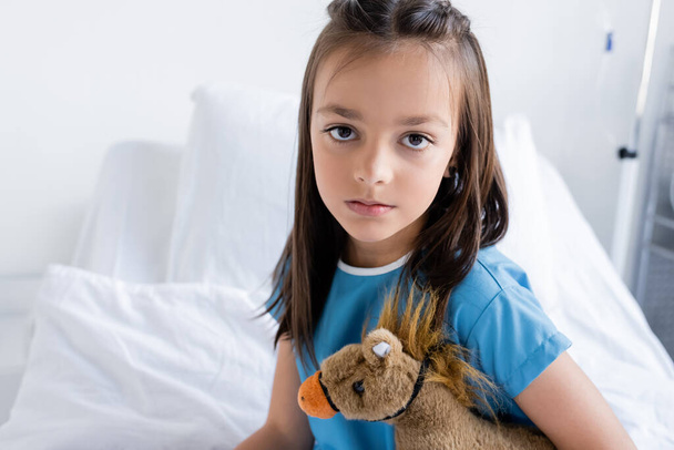 Kind im Patientenkleid hält Spielzeug in der Hand und blickt in Kamera auf verschwommenem Krankenhausbett  - Foto, Bild