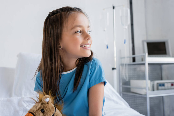 Pozitivní dítě v pacientské róbě dívá pryč v blízkosti měkké hračky na posteli na klinice  - Fotografie, Obrázek
