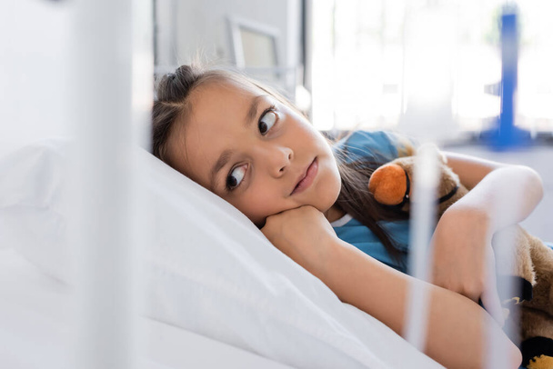 Ребенок обнимает мягкую игрушку, когда лежит в палате больницы  - Фото, изображение