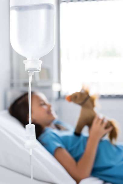 Garrafa de terapia intravenosa perto de criança borrada com brinquedo na cama na clínica  - Foto, Imagem