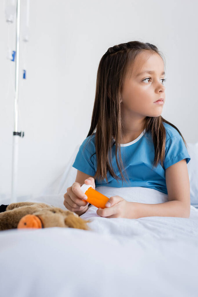 Kid in patient gown holding pills on bed in hospital  - Photo, Image
