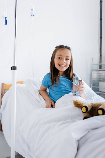 Smiling girl in patient gown holding glass of water on bed in hospital  - Photo, Image