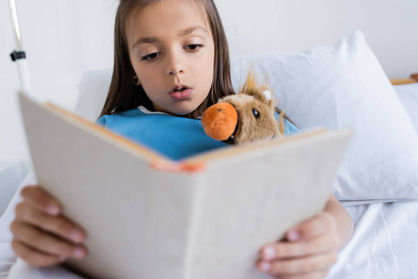 Kid in patient gown reading book near toy on hospital bed  - Foto, Imagen