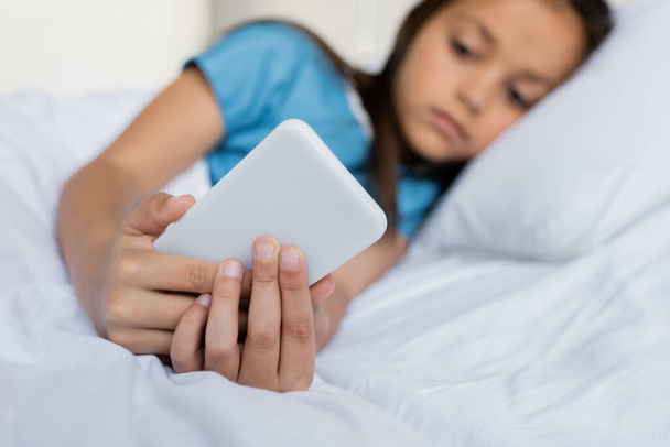 Niño borroso usando el teléfono celular mientras está acostado en la cama del hospital  - Foto, Imagen