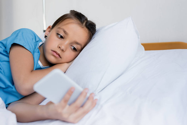 Kid in patient gown using blurred smartphone on hospital bed  - Φωτογραφία, εικόνα