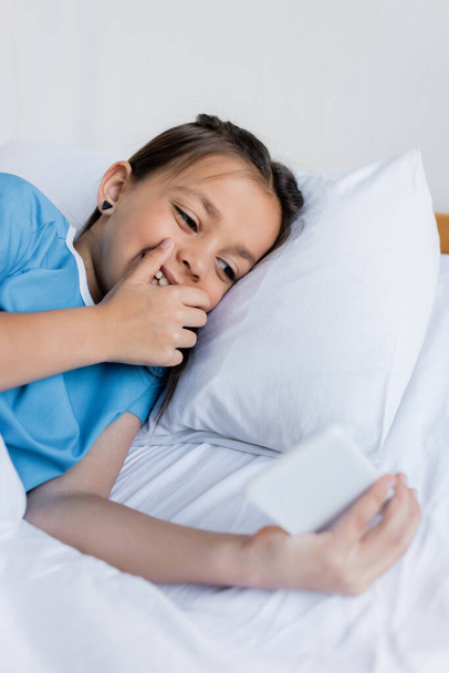 Wesoły dzieciak korzystający z rozmytego telefonu komórkowego leżącego na łóżku w szpitalu  - Zdjęcie, obraz