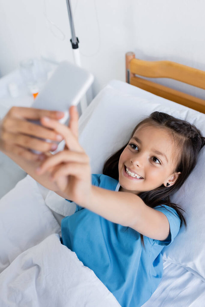 High angle view of smiling kid taking selfie on smartphone in hospital ward  - Photo, Image