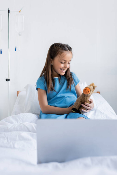 Cheerful kid holding toy near blurred laptop on hospital bed  - Фото, изображение