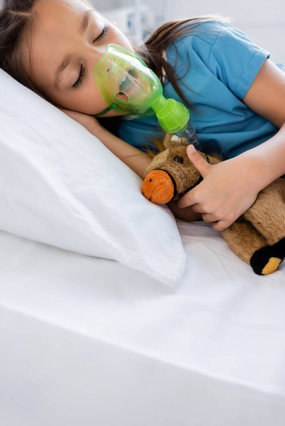Hasta önlüklü ve oksijen maskeli bir kız hastanedeki yatakta oyuncak tutuyor.  - Fotoğraf, Görsel