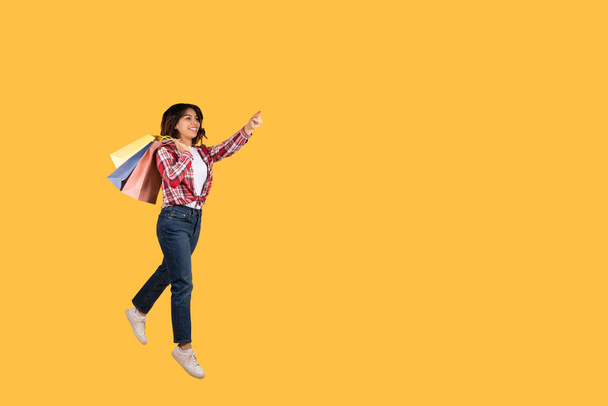 Vrolijke mooie jonge arabische vrouw in casual outfit met kleurrijke boodschappentassen springen of rennen in de lucht over gele studio achtergrond, wijzen op kopieerruimte voor reclame, full length shot - Foto, afbeelding