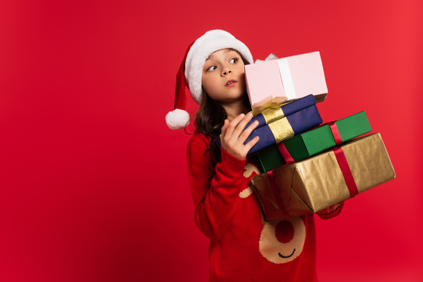 surprised girl in santa hat and sweater holding bunch of Christmas presents isolated on red  - Φωτογραφία, εικόνα