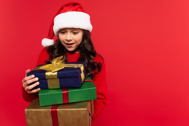happy girl in santa hat and sweater holding bunch of Christmas presents isolated on red - Photo, Image