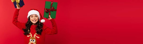 Ευτυχισμένο κορίτσι σε καπέλο και πουλόβερ Σάντα εκμετάλλευση χριστουγεννιάτικα δώρα που απομονώνονται σε κόκκινο, banner - Φωτογραφία, εικόνα