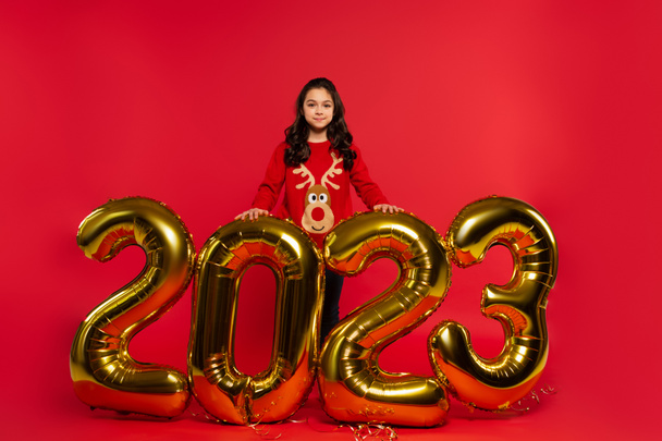 full length of happy girl in sweater standing near balloons with 2023 numbers on red - 写真・画像