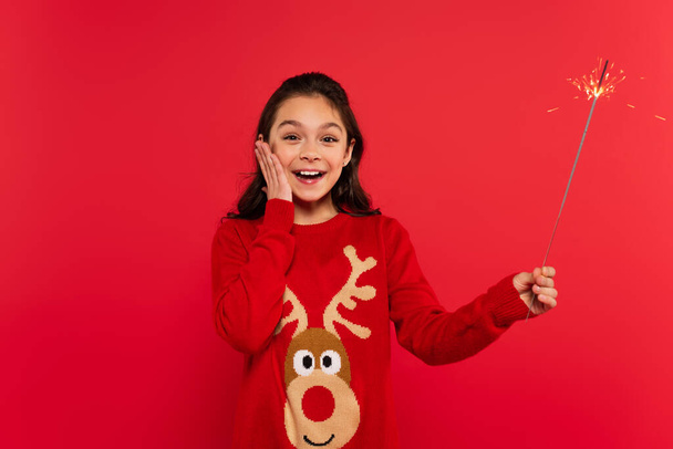 happy girl in Christmas sweater holding shiny sparkler isolated on red - Foto, Imagem