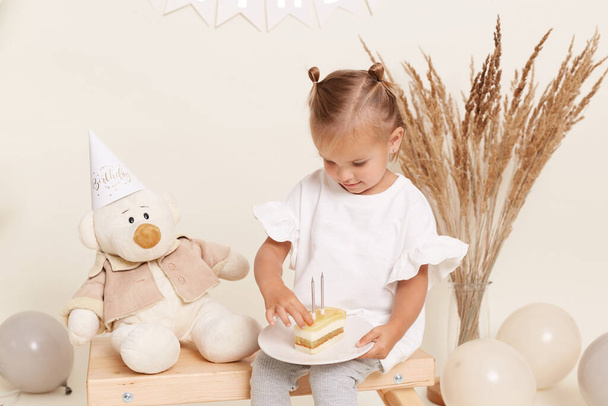 Portrait of blonde child celebrating birthday, toddler with two small funny hair buns sitting with cake and candles with teddy bear nearby and surrounded with balloons, festive atmosphere, party. - Photo, Image