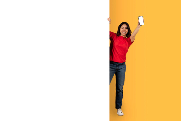 Cool middle eastern young woman in casual outfit student standing by empty white advertising board and showing brand new smartphone with blank screen, yellow studio background, mockup, copy space - Photo, Image