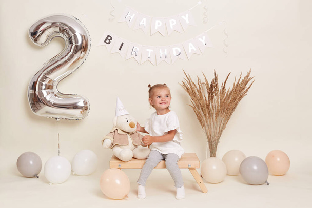 Portrait of little cute blond girl sitting on wooden bench among balloons, celebrating two years, posing in festive room with decoration and big number two made of inflatable foil balloon. - Foto, imagen