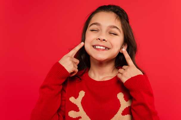 cheerful child in winter sweater pointing with fingers at face isolated on red - Foto, immagini