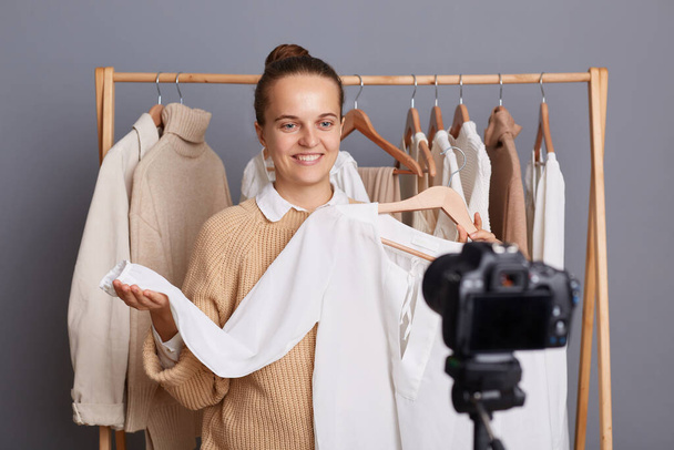 Portrait of happy delighted smiling woman standing near hanger rack with attires in fashion store or at home in wardrobe, showing white blouse to her followers, selling outfits. - Foto, imagen