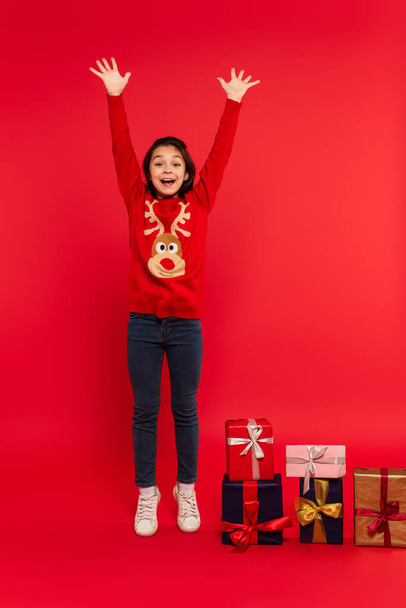 full length of excited child in knitted sweater gesturing near Christmas presents on red - Zdjęcie, obraz