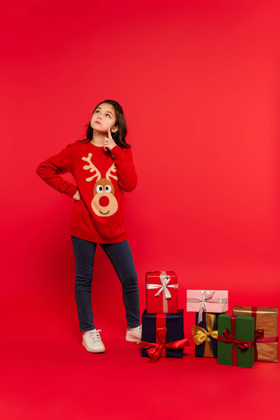 longitud completa de niño pensativo en suéter de punto posando con la mano en la cadera cerca de regalos de Navidad en rojo - Foto, Imagen