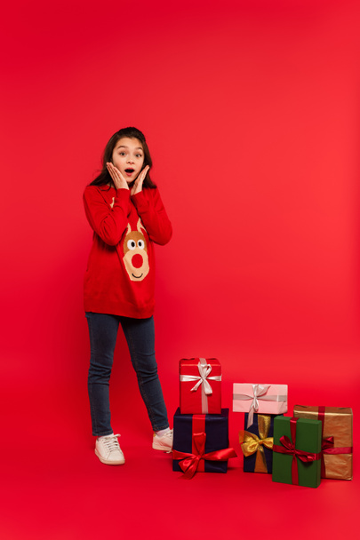 full length of shocked child in knitted sweater near Christmas presents on red - Fotó, kép