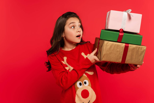 amazed girl in winter sweater holding bunch of Christmas presents and smiling isolated on red - Φωτογραφία, εικόνα