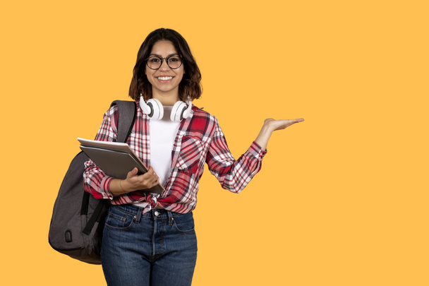 Señorita árabe joven atractiva positiva en estudiante casual que muestra espacio de copia en el fondo del estudio amarillo, con gafas, llevando mochila, sosteniendo el ordenador portátil y libros. Educación, escuela, universidad - Foto, Imagen