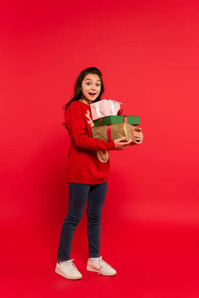 full length of happy girl in winter sweater holding bunch of Christmas presents and smiling on red - Fotó, kép