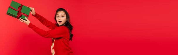 shocked girl in winter sweater holding wrapped Christmas present isolated on red, banner - Fotoğraf, Görsel