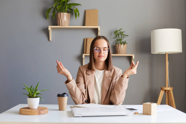Portrait of attractive woman sitting at workplace with laptop and raising hands in mudra gesture, meditating resting at home office, posing with closed eyes. - Фото, изображение