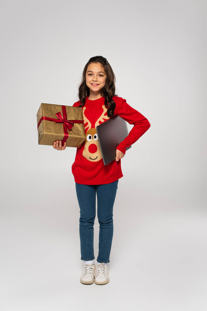 full length of happy girl in red sweater holding laptop and Christmas present on grey - Foto, afbeelding