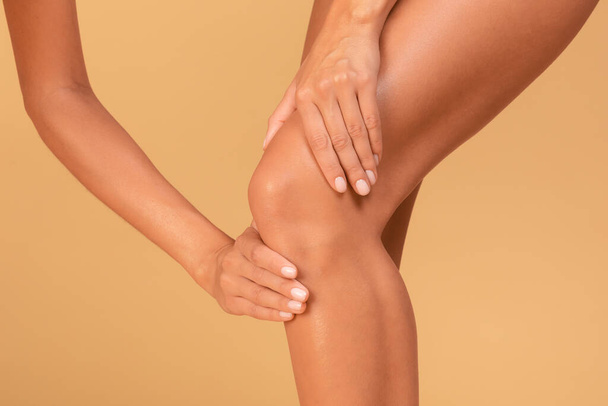 Legs problems. Young caucasian woman touching her inflamed knee with acute pain, standing over beige studio background, closeup, cropped - 写真・画像