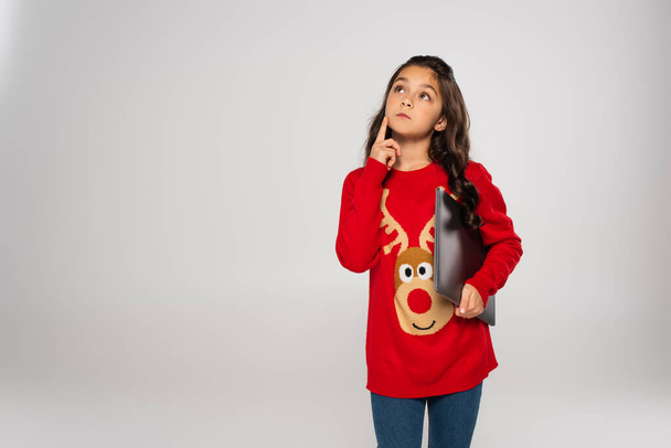 Nachdenkliches Kind im roten Weihnachtspullover hält Laptop isoliert auf grau - Foto, Bild