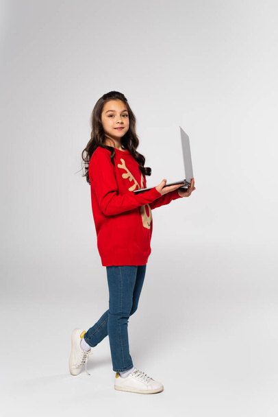 full length of smiling child in red Christmas sweater holding laptop on grey - 写真・画像