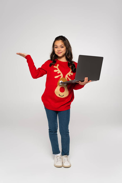 volledige lengte van vrolijk kind in rode trui met laptop en wijzend met de hand op grijs - Foto, afbeelding