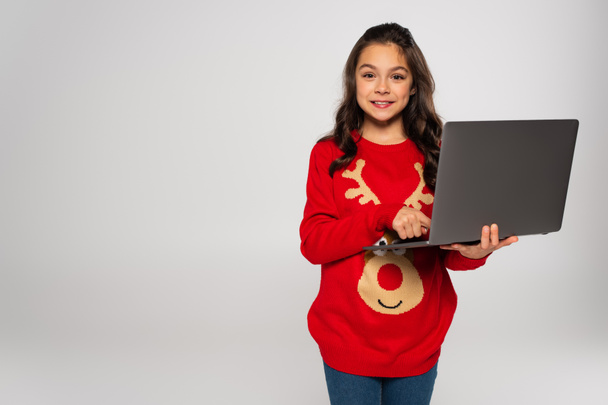 smiling child in red Christmas sweater using laptop isolated on grey - Foto, afbeelding