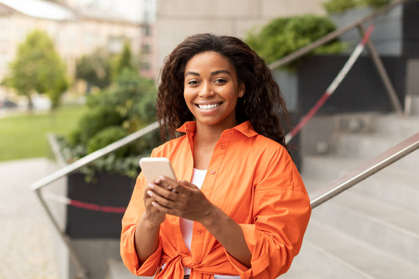 Smiling millennial black female typing on smartphone, chatting and reading blog, enjoys walk and look at camera in city. Modern mobile device and application, great offer and ad, social networks - Photo, Image