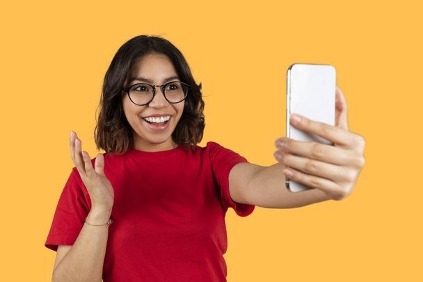 Leuke jonge arabische vrouw in rood t-shirt met een bril op met behulp van gloednieuwe smartphone, kijken naar mobiele telefoon scherm, glimlachen en gebaren, selfie of het hebben van online partij, gele achtergrond - Foto, afbeelding