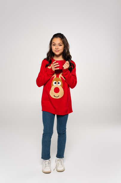 πλήρες μήκος του ευτυχισμένου κοριτσιού σε κόκκινο πουλόβερ κρατώντας φλιτζάνι κακάο σε γκρι - Φωτογραφία, εικόνα