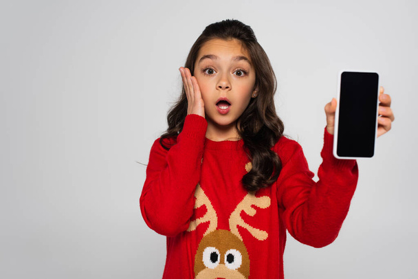 Schockiertes Mädchen im Weihnachtspulli hält Smartphone mit leerem Bildschirm isoliert auf grau  - Foto, Bild