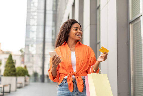 Satisfied happy young black woman with smartphone and packages with purchases and credit card enjoys shopping, looks at shop window in city. Huge sale and modern financial app, huge sale, ad and offer - Foto, Bild