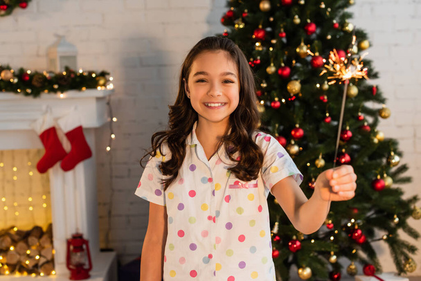 Chica sonriente en pijama sosteniendo chispa y mirando a la cámara durante la celebración de Navidad en casa  - Foto, imagen