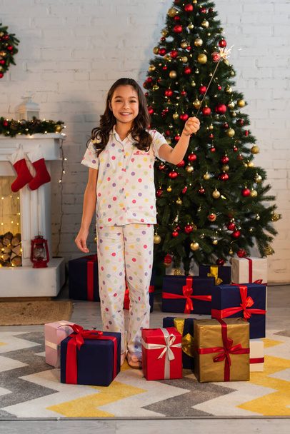 Smiling kid in pajama holding sparkler near gifts and Christmas tree at home  - Фото, зображення