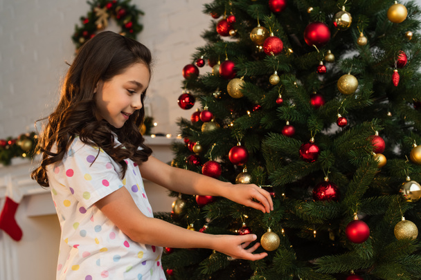 Side view of girl in pajama holding ball near Christmas tree at home  - Фото, зображення